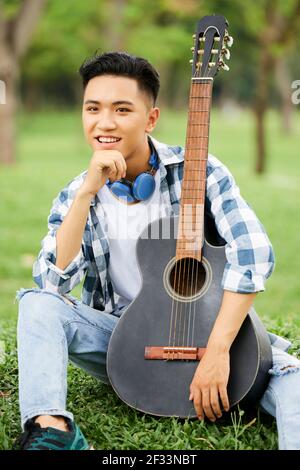 Asian man sitting on green grass with guitar and smiling at camera outdoors Stock Photo