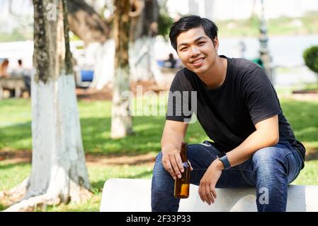 Portrait of Asian young man smiling at camera while drinking beer on the bench in the park Stock Photo