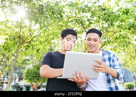 Two Asian men holding laptop and using it together in the city outdoors Stock Photo