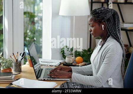 Serious young Black woman with dreadlocks working on laptop at her desk at home and writing essay for colleage Stock Photo