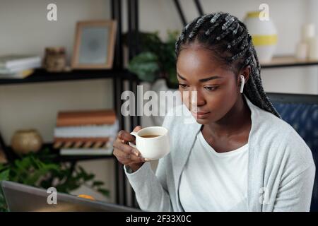 Pretty serious young Black businesswoman drinking morning coffee and reading e-mails on laptop screen Stock Photo