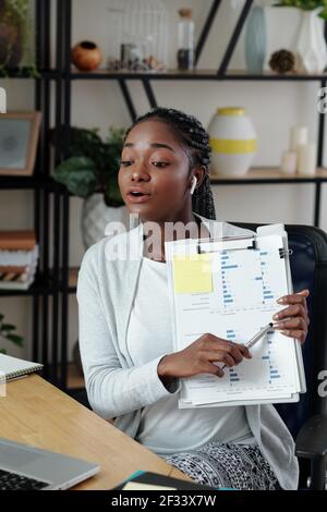 Young female entrepreneur pointng at document with column chart when having online meeting with colleague Stock Photo