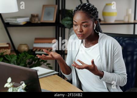 Pretty young serious female entrepreneur wearing earbuds when talking to colleague at online meeting Stock Photo