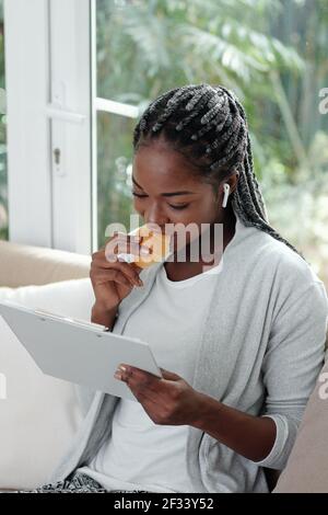 Young Black woman with braided hair eating croissant when reading article in document on clipboard Stock Photo