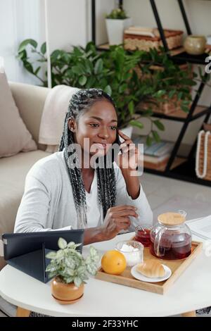 Smiling pretty young Black woman drinking tea with croissant at home and talking on phone with friend Stock Photo