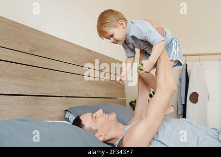 Young man lying on bed and raising his child up they playing in the bedroom Stock Photo