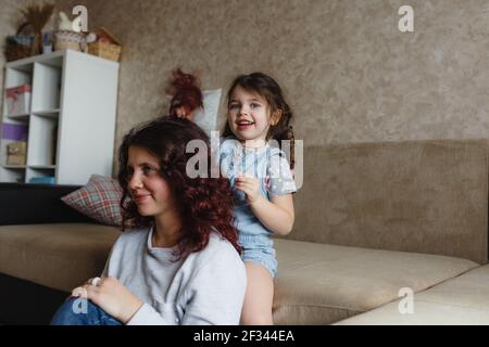 a little girl plays at home with her mother doing her hair while combing her mother s long hair Stock Photo