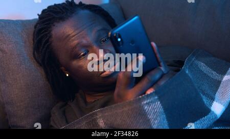 Bored black man relaxing on the couch wrapped in the blanket and using smartphone. High quality photo Stock Photo