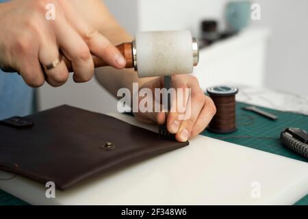 Mens hand holding a leather carving hammer and steel chisel hole punch and makes a leather wallet in his workshop. Working process with a brown Stock Photo