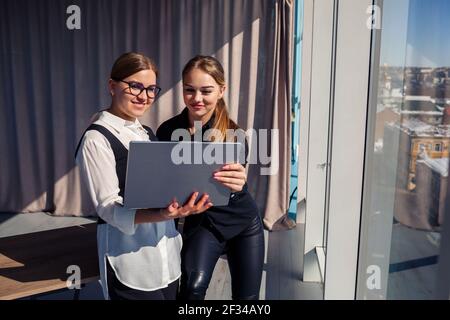 Two successful female architects are talking about a joint project while standing at the window with a laptop. Young women economists dressed in forma Stock Photo
