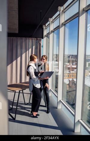 Two successful female architects are talking about a joint project while standing at the window with a laptop. Young women economists dressed in forma Stock Photo