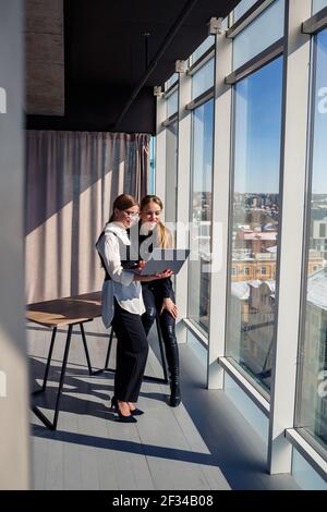 Two successful female architects are talking about a joint project while standing at the window with a laptop. Young women economists dressed in forma Stock Photo