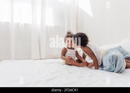 Brother and sister African Americans play together on white bed in loft interior. Siblings having fun among the blue pillows in the morning. Boy kissi