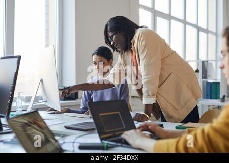 Side view portrait of female team leader instructing colleague and pointing at screen while working with multi-ethnic team of software developers in o Stock Photo