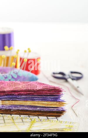 Ruler, cutting mat, stack of multi-colored pieces of fabric, pincushion, spools of thread, scissors, needle and thread on a white surface. Space for t Stock Photo