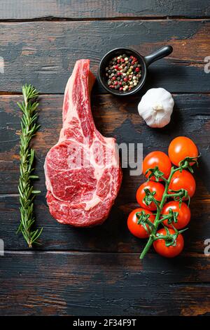 Raw uncooked black angus Bone In Steak in rustic style on wooden table background. with tomato garlic herbs and peppercorns rosemary close up Stock Photo