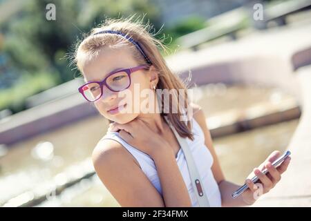 Young pre-teen blonde girl holds mobile phone in her hand sitting on the edge of a fountain on a sunny summer day. Stock Photo