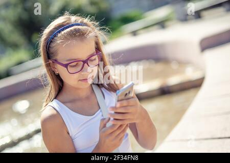 Young pre-teen blonde girl looks into the mobile phone in her hand sitting on the edge of a fountain on a sunny summer day. Stock Photo