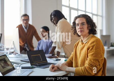 Portrait of young long haired man looking at camera while using laptop in office with diverse team of software developers, copy space Stock Photo
