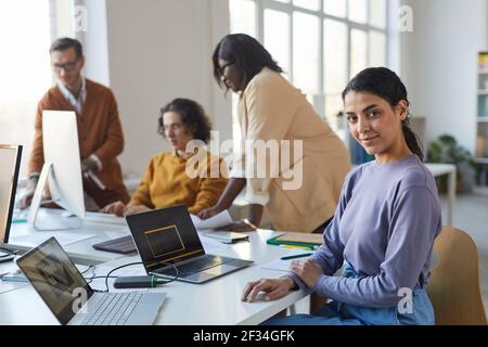 Portrait of young Indian woman looking at camera while using laptop in office with diverse team of software developers, copy space Stock Photo