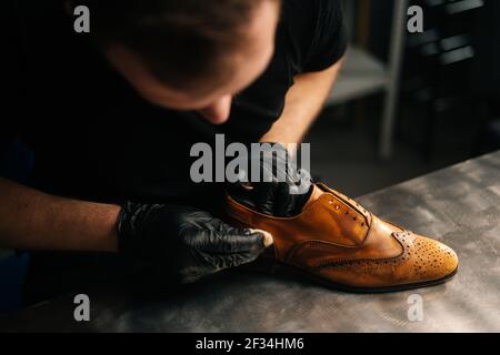 Top close-up view of professional shoemaker wearing black gloves polishing old light brown leather shoes.  Stock Photo