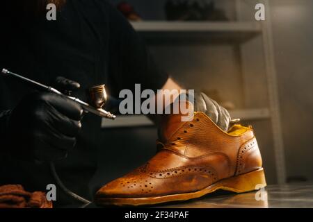 Close-up side view of unrecognizable shoemaker wearing black gloves spraying paint of light brown leather shoes. Stock Photo