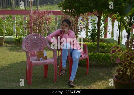 Close up of Indian Bengali teenage girl wearing jeans and pink upper sitting on a chair in the lawn hands on chin smiling, selective focusing Stock Photo