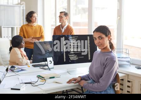 Portrait of female IT developer looking at camera while posing against computer with code on screen in software production studio, copy space Stock Photo