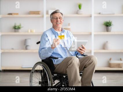 Happy disabled elderly man in wheelchair shopping online with tablet and credit card, indoors Stock Photo
