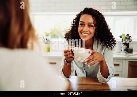 Mixed race female wiith afro drinking coffee with caucasian friend in kitchen smiling Stock Photo