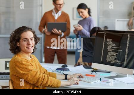 Portrait of young IT developer looking at camera and smiling while using computer with code on screen in software production studio, copy space Stock Photo