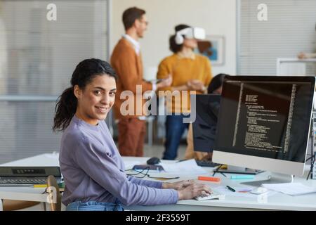 Portrait of female IT developer looking at camera and smiling while using computer with code on screen in software production studio, copy space Stock Photo