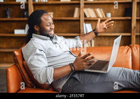 Happy young African American man with dreadlocks in casual attire lying down on couch, talking with emotions, gesturing, video-calling online with laptop on lap, chatting with family, friend on webcam Stock Photo