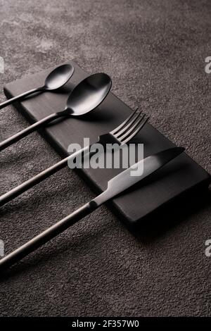 Black matte cutlery set-fork, spoon, knife, on dark concrete. Minimalistic still life, stylish tableware. vertical photo close-up. Stock Photo