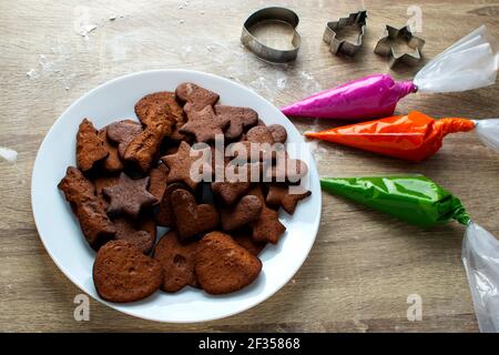 On a wooden table there is a white plate on which gingerbread cookies and icing next to them Stock Photo