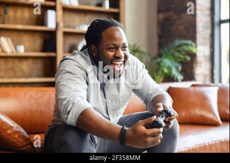 Portrait of young African American guy sitting on the couch in the living room, holding joystick, screaming, and winning a video game match, male addicted gamer with an open mouth is about to succeed Stock Photo