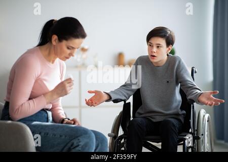 Conflict between parent and teenager. Sad mother crying, teen son in wheelchair making excuses or apologizing at home Stock Photo