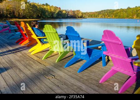 Man relaxing in a row of brightly colored adirondack chairs along waterfront in fall Stock Photo