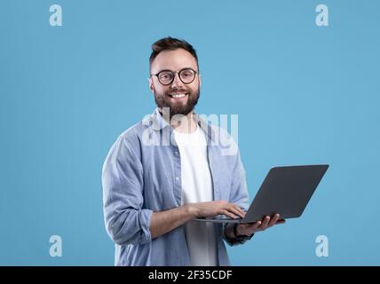 Cheerful millennial guy in glasses using laptop computer for online work or communication on blue studio background Stock Photo