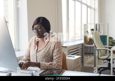 Portrait of young African-American woman using computer while working at desk alone in modern white office, copy space Stock Photo