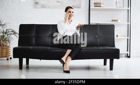 smiling businesswoman pointing with hand while talking on smartphone on sofa Stock Photo