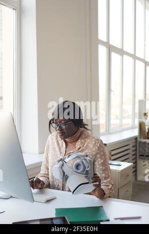 Vertical portrait of young African-American woman using computer while working at desk alone in modern white office, copy space Stock Photo