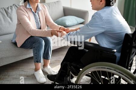 Cropped view of disabled tenager in wheelchair holding hands with female psychologist, closeup Stock Photo