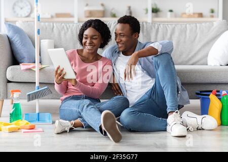 Black couple with cleaning tools resting, using digital tablet Stock Photo