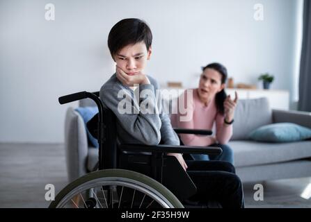 Unhappy teen boy in wheelchair having conflict with his mother, listening to her scolding at home. Relationship problems Stock Photo