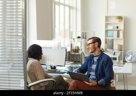 Side view portrait of smiling contemporary businessman talking to African-American colleague while using laptop in white office, copy space Stock Photo