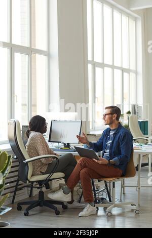 Vertical full length portrait of smiling contemporary businessman talking to African-American colleague while using computer in white office, copy spa Stock Photo