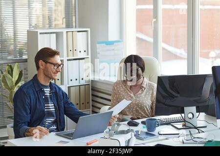 Portrait of smiling contemporary businessman talking to African-American colleague while using computers in white office interior Stock Photo