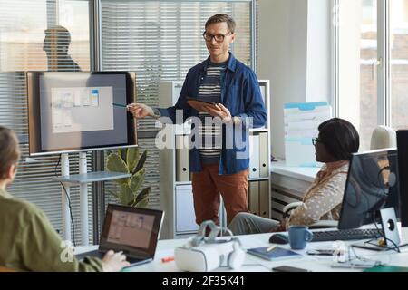 Portrait of contemporary businessman pointing at digital screen while giving presentation at team meeting in software production office, copy space Stock Photo