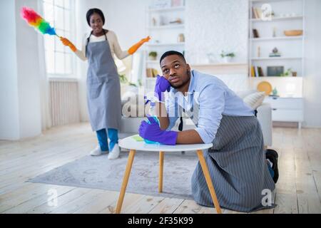 Young black guy feeling tired from housework, his angry wife scolding him for doing nothing, copy space Stock Photo